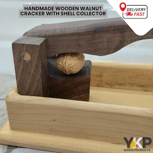 Può includere: Schiaccianoci in legno di noce fatto a mano con un raccoglitore di gusci. Lo schiaccianoci è in legno scuro e ha una base in legno chiaro. Una noce viene schiacciata nel dispositivo.