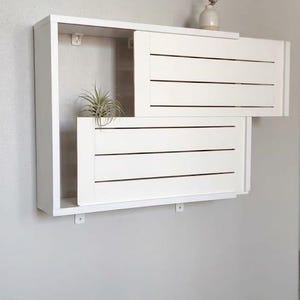 May include: A white wall-mounted shelf unit featuring two sliding panels with horizontal slats. The unit has an open rectangular frame and includes a small air plant inside the left section and a small vase with a dried stem on the top right.