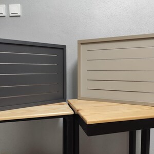 Può includere: Due pannelli rettangolari orizzontali con un design a doghe. Uno è grigio scuro, l'altro beige. Entrambi sono incorniciati e poggiano su tavoli di legno con gambe in metallo nero. Lo sfondo è una parete grigio chiaro.