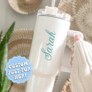 May include: White travel mug with a teal blue inscription that says "Sarah". The mug has a lid with a straw hole and a handle. A sticker on the mug says "Custom Just For Her!"