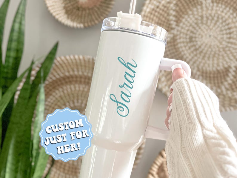 May include: White travel mug with a teal blue inscription that says "Sarah". The mug has a lid with a straw hole and a handle. A sticker on the mug says "Custom Just For Her!"