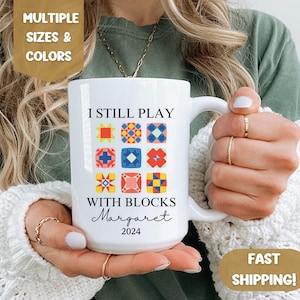 May include: White ceramic mug with a colorful quilt block design and the text "I still play with blocks Margaret 2024".