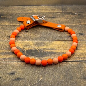 Op de afbeelding: Hondenhalsband met oranje en perzik kleurige kralen, met een oranje leren gesp en riem. De gesp is van zilverkleurig metaal. De kralen zijn in een cirkelvorm gerangschikt.