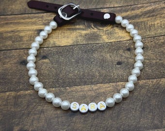 Puede incluir: Un collar de perlas con el nombre "SARA" escrito con cuentas de letras blancas con letras doradas. El collar tiene una correa de cuero burdeos oscuro con una hebilla plateada.