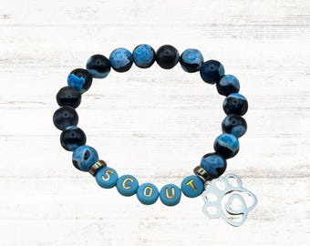 Könnte beinhalten: Ein Armband aus blauen und schwarzen Achatperlen mit einem silbernen Pfotenabdruck-Anhänger und dem Namen "SCOUT" in goldenen Buchstaben auf blauen Perlen.