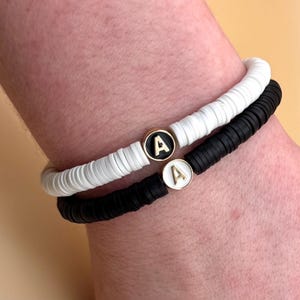 Puede incluir: Dos pulseras con cuentas blancas y negras. Cada pulsera tiene un dije dorado con la letra "A".