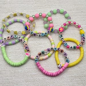 Puede incluir: Un conjunto de seis pulseras de cuentas coloridas con diferentes colores pastel y palabras como "Hoppy Easter", "Eggstra", "Sweet as Candy" y "Some Loves You".