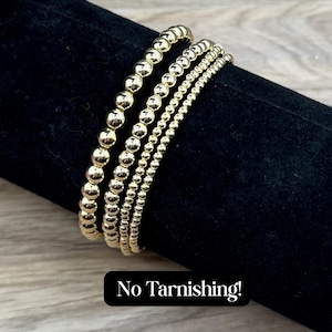 May include: Four gold beaded bracelets stacked on a black surface. The text "No Tarnishing!" is written below the bracelets.