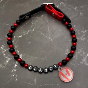 Könnte beinhalten: Halsband für Hunde mit schwarzen und roten Perlen, mit dem Namen "CLANCY" in weißen Buchstabenperlen geschrieben. Das Halsband hat einen schwarzen Lederriemen und einen roten Akzent. Ein runder, silberner Anhänger mit rotem Logo ist angebracht.
