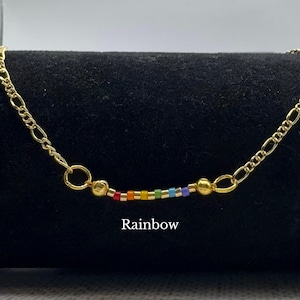 May include: Gold-colored necklace with a rainbow bead design. The necklace features a chain with a series of small, colorful beads in a rainbow pattern. The word "Rainbow" is printed below the necklace.