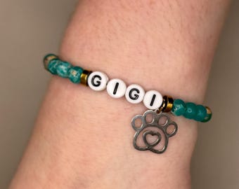 Könnte beinhalten: Ein grünes und goldenes Perlenarmband mit weißen Buchstabenperlen, die "GIGI" buchstabieren, und einem silbernen Pfotenabdruck-Anhänger.