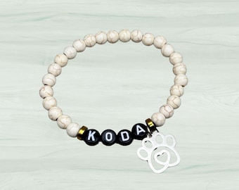 Könnte beinhalten: Weißes Perlenarmband mit einem schwarzen Pfotenabdruck-Anhänger und dem Namen "KODA" in schwarzen Perlen.