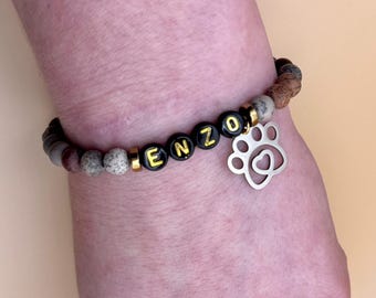 Könnte beinhalten: Ein Armband mit braunen und grauen Perlen, schwarzen Buchstabenperlen, die "ENZO" buchstabieren, und einem silbernen Pfotenabdruck-Anhänger mit einem Herz darin.