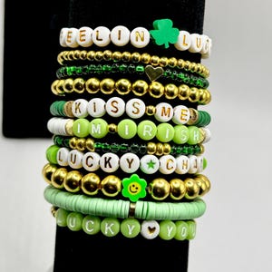 Könnte beinhalten: Ein Stapel Armbänder mit Perlen in Grün- und Goldtönen. Einige Armbänder haben weiße Buchstabenperlen mit Aufschriften wie "FEELIN LUCKY", "KISS ME I'M IRISH" und "LUCKY YOU". Zu den Charms gehören ein Kleeblatt und ein Smiley.