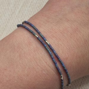 May include: Two beaded bracelets on a wrist. The bracelets are made of small, iridescent blue and purple beads with silver accents.