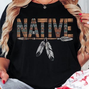 May include: Black t-shirt with the word "NATIVE" in a decorative, patterned font. Below the word are two arrows and two white feathers. The shirt is a casual style.