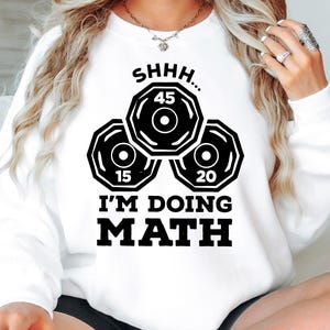 May include: White sweatshirt with a black graphic. The graphic features three weight plates with the numbers 15, 20, and 45, and the text "SHHH... I'M DOING MATH" in bold letters. The sweatshirt is a casual style.