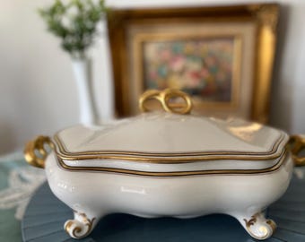 May include: A white porcelain serving dish with a gold trim and a lid. The dish has four feet and is sitting on a blue and white patterned plate.