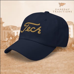 以下が含まれることがあります： ゴールドの筆記体で「Tech」と刺繍されたネイビーブルーの野球帽。 つばがカーブしており、クラシックな6パネルデザインです。「Gameday Traditions」のロゴが右上に見えます。