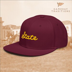 May include: A maroon baseball cap with a flat peak. The word "State" is embroidered in yellow script across the front. The cap is from "Gameday Traditions".
