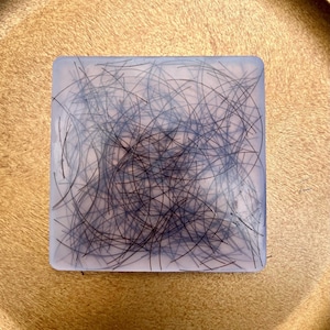 Op de afbeelding: Een vierkant, transparant blok van hars met ingebedde zwarte haarstrengen. De haarstrengen zijn willekeurig gerangschikt en creëren een draaiend patroon.