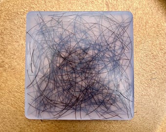 May include: A clear square block of resin with black hair strands embedded inside. The hair strands are randomly arranged and create a swirling pattern.