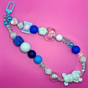 May include: A blue and white beaded keychain with a variety of charms, including a paw print, a heart, a candy bar, and a bear. The keychain is on a pink background.