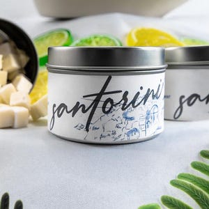 May include: Two silver candle tins with white labels. The labels have the word "santorini" written in black cursive. The tin on the left has a blue and white illustration of a town on the label.