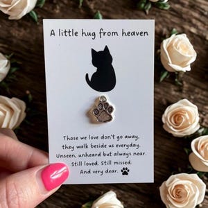 May include: A silver-toned metal paw print charm attached to a white rectangular card. The card features a black silhouette of a cat and the text: A little hug from heaven. Those we love don't go away, they walk beside us everyday. Unseen, unheard but always near. Still loved. Still missed. And very dear.
