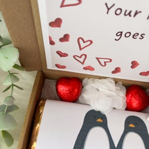 May include: A Valentine's Day gift box with a printed card featuring two penguins. The box contains two heart-shaped chocolates wrapped in red foil, a gold-wrapped chocolate bar, and white tissue paper. The card has red hearts.