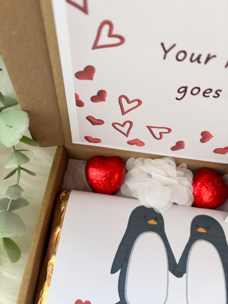 May include: A Valentine's Day gift box with a printed card featuring two penguins. The box contains two heart-shaped chocolates wrapped in red foil, a gold-wrapped chocolate bar, and white tissue paper. The card has red hearts.