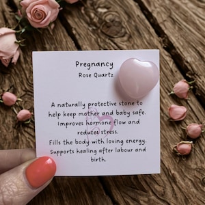 May include: A heart-shaped rose quartz stone with the word "Pregnancy" on a white card. The card also includes text about the stone's benefits. Pink roses and buds are scattered around the card on a wooden surface.
