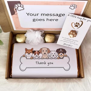 May include: A cardboard gift box containing a chocolate bar wrapped in white paper with a cartoon dog illustration and the text Thank you, two gold-wrapped chocolates, a white fabric flower, and a card with a paw print charm and text.