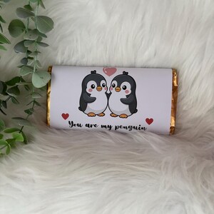May include: A chocolate bar wrapped in gold foil with a white label featuring two cartoon penguins. The penguins are black, white, and orange, with a pink heart above them. The text "You are my penguin" is below the penguins.