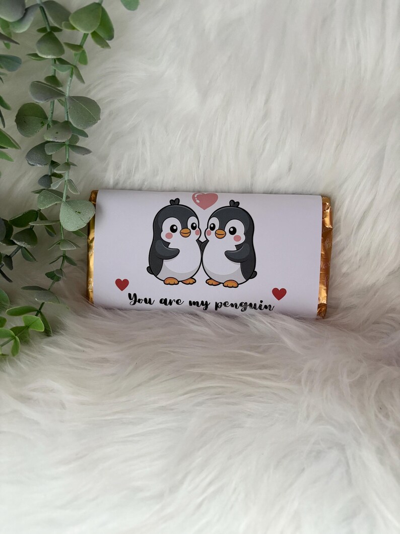 May include: A chocolate bar wrapped in gold foil with a white label featuring two cartoon penguins. The penguins are black, white, and orange, with a pink heart above them. The text "You are my penguin" is below the penguins.
