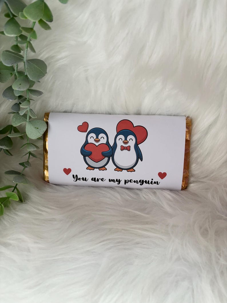 May include: A chocolate bar wrapped in gold foil with a white paper label. The label features two cartoon penguins holding red hearts, with the text "You are my penguin" and small red hearts.