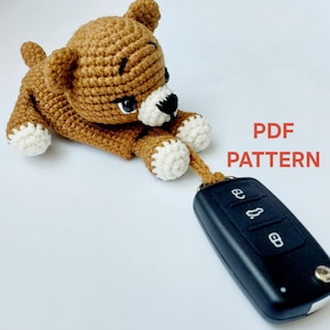 Op de afbeelding: Een gehaakte bruine beren sleutelhanger met witte accenten op de poten en snuit, die een zwarte autosleutel vasthoudt. De tekst "PDF PATTERN" staat in het rood. Een handgemaakt item.