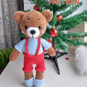 May include: A crocheted brown teddy bear wearing a blue shirt and red overalls. The bear is standing in front of a small artificial Christmas tree with red ornaments. The text "PDF PATTERN" is written in red letters on the tree.