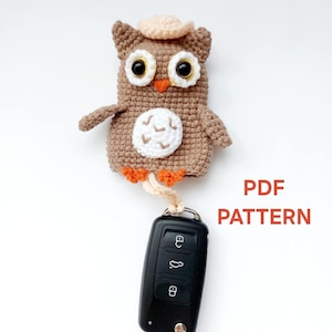 Può includere: Coprichiave a forma di gufo all'uncinetto, marrone con cappello beige e dettagli bianchi. Il gufo ha grandi occhi rotondi e piedi arancioni. È attaccata una chiave per auto nera. Il testo sull'immagine recita "PDF PATTERN".