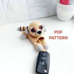 May include: A tan crochet lemur keychain with gray accents and a black nose. The lemur is attached to a black car key. The text "PDF PATTERN" is displayed in red.