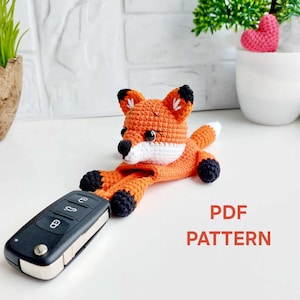 Pode incluir: Raposa laranja de crochê com detalhes em preto e branco, segurando uma chave do carro. A raposa tem o peito e a ponta da cauda brancos. O texto "PDF PATTERN" está em vermelho. A raposa está em uma superfície branca.