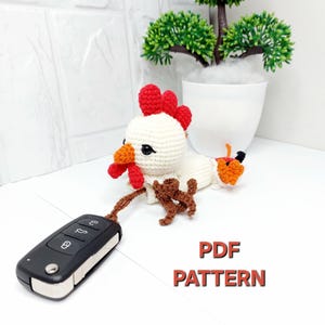 May include: A crocheted chicken keychain with a white body, red comb and wattle, and orange feet, attached to a black car key fob. The text PDF PATTERN is printed in red and black on the white surface next to the crocheted chicken figure.