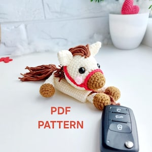 May include: A handmade crochet horse in white with brown mane and tail, and brown hooves. It has a red bridle and is next to a car key. The text "PDF PATTERN" is displayed below the horse.