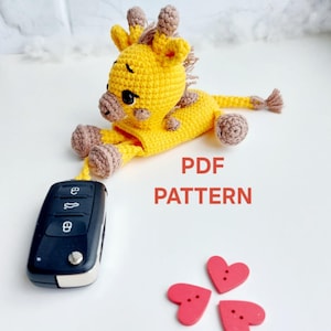 May include: A handmade yellow crochet giraffe toy with brown spots and accents. The giraffe is lying down next to a black car key fob and three red heart-shaped buttons. The text "PDF PATTERN" is in red.