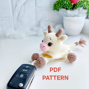May include: A cream-colored crocheted cow amigurumi with brown hooves, horns, and tail, and a pink nose. The cow is lying down next to a car key. The text "PDF PATTERN" is in red.