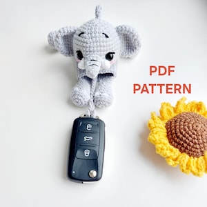 Op de afbeelding: Een gehaakte grijze olifant sleutelhanger met een zwarte autosleutel eraan. De olifant heeft grote oren en een slurf. Een gehaakte gele en bruine zonnebloem staat in de rechter benedenhoek. De tekst "PDF PATTERN" staat in de afbeelding.
