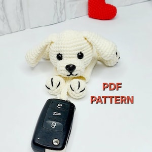 Peut inclure: Un protège-clés en forme de chiot crocheté de couleur crème, avec des yeux et un nez noirs, tenant une clé de voiture noire. Un petit cœur rouge crocheté est en arrière-plan. Le texte "PDF PATTERN" est en rouge.