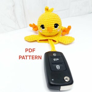 May include: A handmade yellow crochet chick key cover with orange accents, holding a black car key. The chick has black eyes and a small orange beak. The text "PDF PATTERN" is displayed in red.