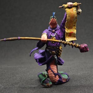 May include: A painted miniature figurine of a snake-like creature with a human-like torso wearing a purple hooded robe. The figure holds a long wooden staff in one hand and a yellowed scroll with red text in the other, featuring a green scaled tail.
