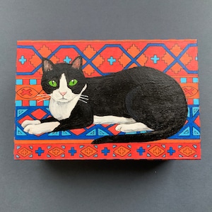 May include: A rectangular wooden box featuring a black and white cat with green eyes, resting on a colorful patterned background. The background has a geometric design in red, orange, blue, and white.